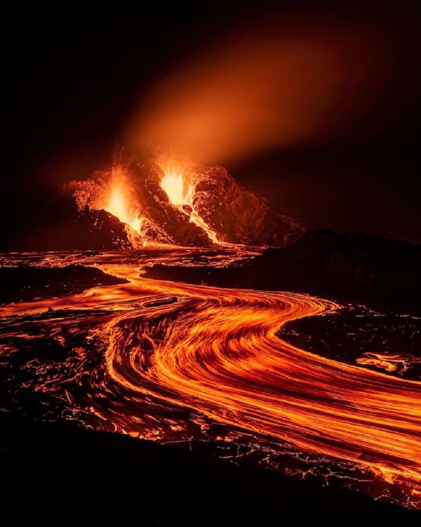 A photograph showing the active eruption from the Reykjanes Peninsular. Pictured here is the new cauldron-like cone forming around one of the main eruptive vents. Snaking away from the cone is a  glowing molten river of lava. 