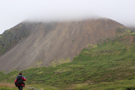 A sandy-reddish coloured, dome-shaped exposure (the Sandfell laccolith) in a steep, grassy slope. A person stands at the base of the image, with clouds covering the peak of the slope.