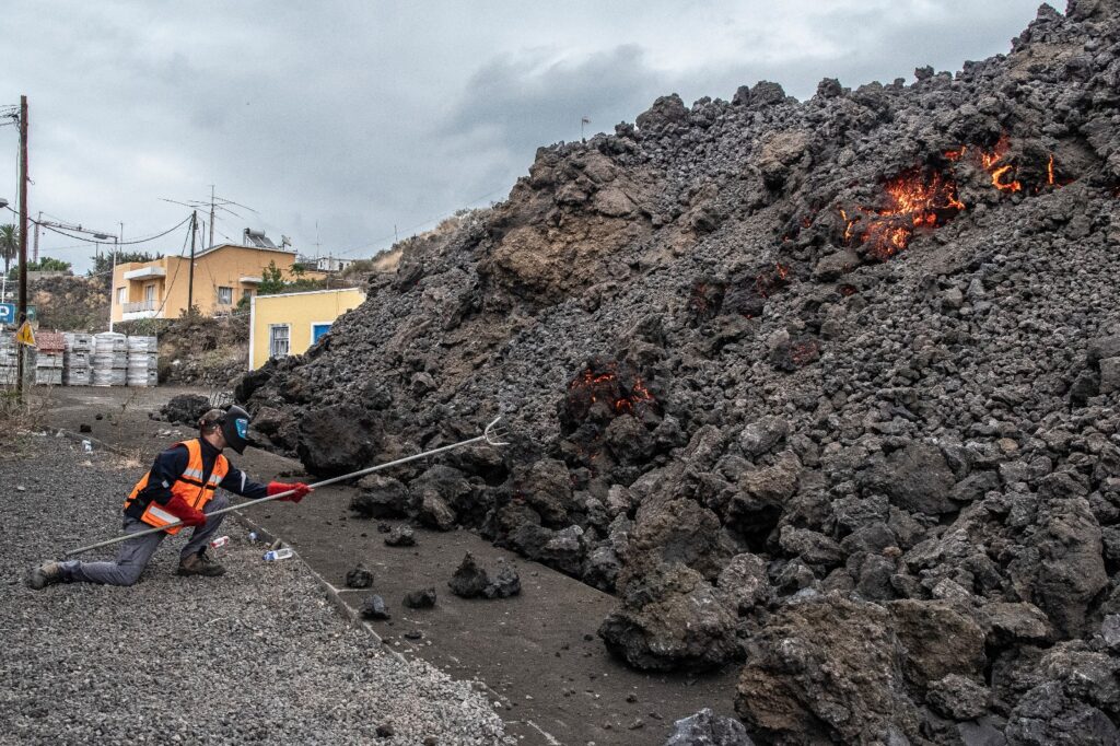 A petrologist sampling an active lava flow. The flow is largely dark grey in colour and has a rubble like appearance. A small portion of glowing orange lava can be seen flowing down the front of the flow. 