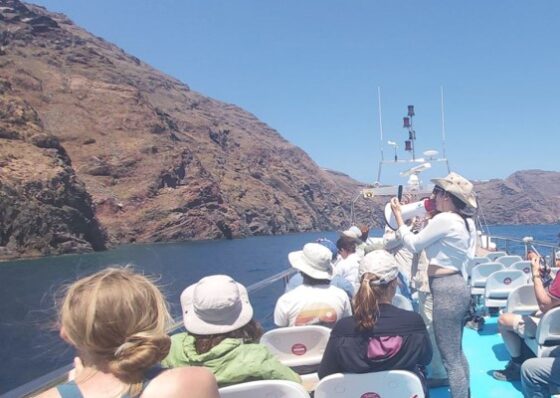 A group of people on a boat looking at an outcrop, listening to a woman using a megaphone.