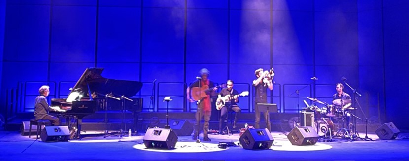 The 5 members of the Vasilis Rokopoulos quintet performing on stage playing their respective instruments (piano, two guitars, drums and trumpet).