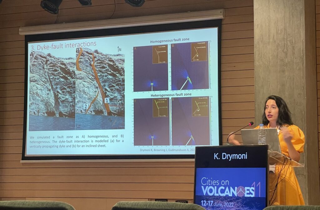 Kyriaki Drymoni stood at the podium presenting her work at COV11, with a slide showing a photo of a dyke-fault interaction outcrop compared with numerical modelling results.