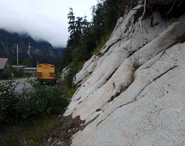 A photograph of a grey rock outcrop on a road cut with trees and steep slopes in the background.